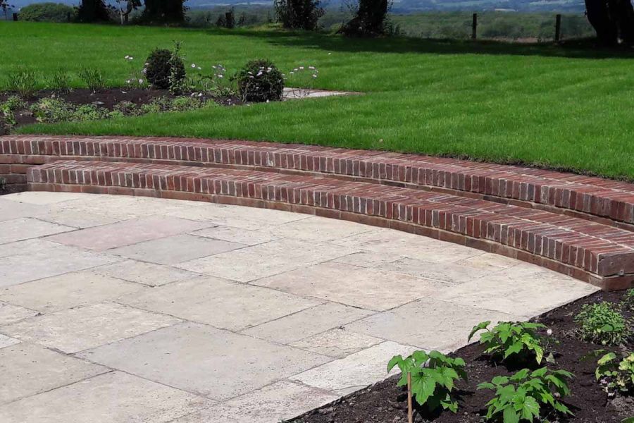 2 wide, shallow, curved steps of Old English garden wall bricks descend from grass to paved area. Design by Arun Landscapes.
