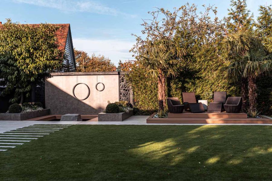Firepit backed by high wall faced with Vulcano Ceniza porcelain cladding in modern garden with raised patio under exotic trees.