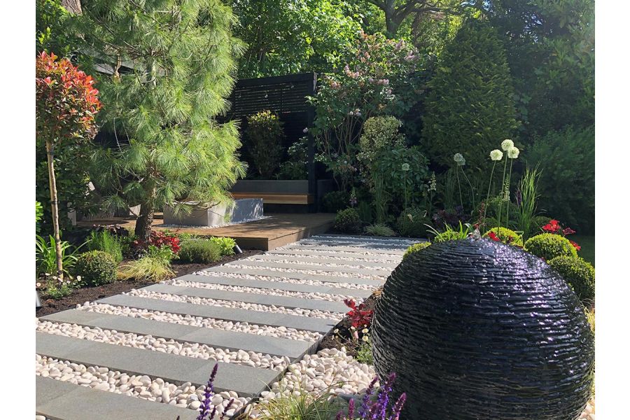 Egg-shaped slate water feature next to striped path of Black granite plank paving and white pebbles and beds with topiary.