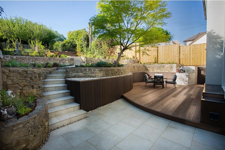 Multi-level garden paved with Gea Porcelain paving and steps, featuring a Millboard deck area with garden furniture on the right.