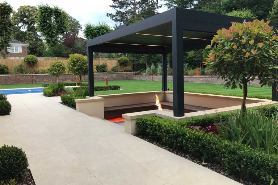 Wide area of Golden Stone Porcelain patio tiles with steps down to walled, sunken pergola-covered seating area with firepit.