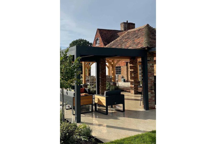 Garden furniture sits underneath dark metal pergola with wet golden stone porcelain paving showing off its texture.