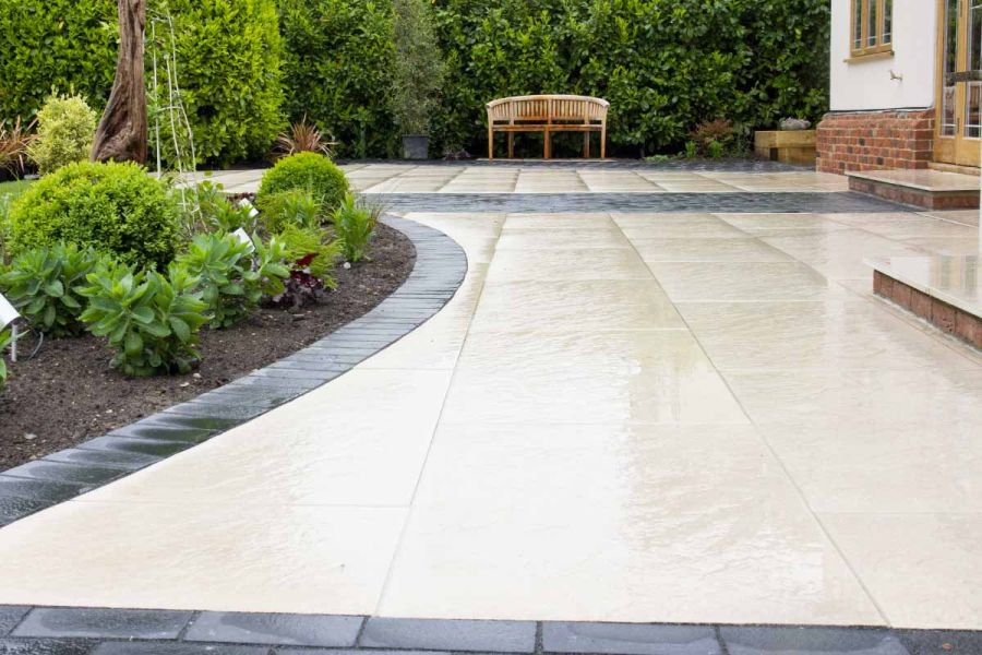 Wide, low shot of Golden Stone Porcelain Paving in the wet, showing off the texture. Flowerbed to the left, wooden bench in the distance.