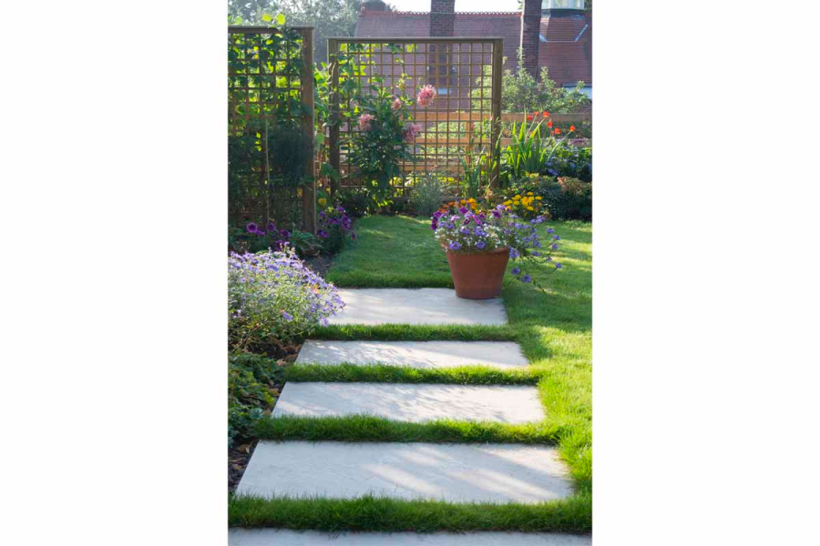 Golden Stone Porcelain paving tiles set into lawn, trellis panels ahead, with flower bed on left and large flower pot with plant.