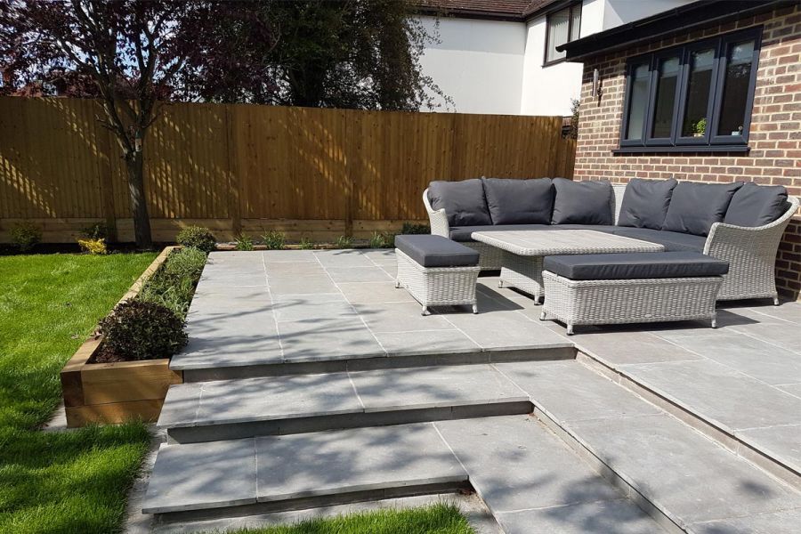 In suburban garden, 3 L-shaped steps descend to lawn from Graphite Grey limestone patio with rattan corner sofa set.