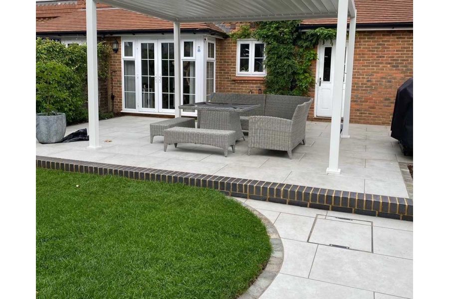 White pergola covers lounge set on brick-edged patio of Florence Grey porcelain slabs by lawn. Design by GRC Landscapes.