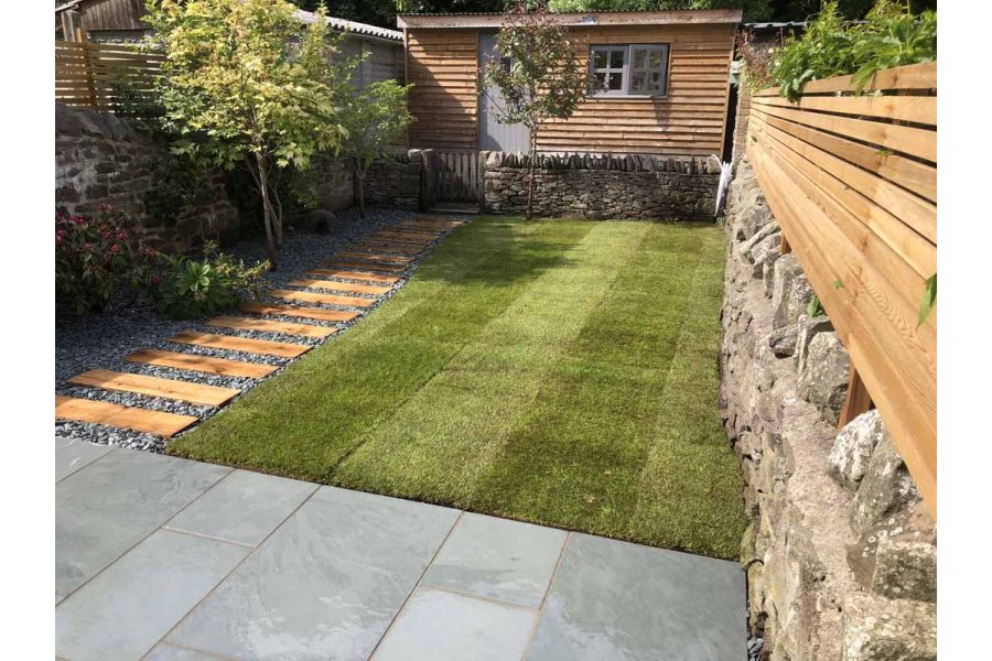 Patio with different widths of Brazilian Grey slate paving slabs fronts garden with lawn and plank path leading to garden office.