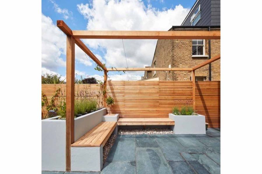 Rectilinear raised beds support wooden bench seating on 2 sides of pergola covered area paved with Green slate paving.