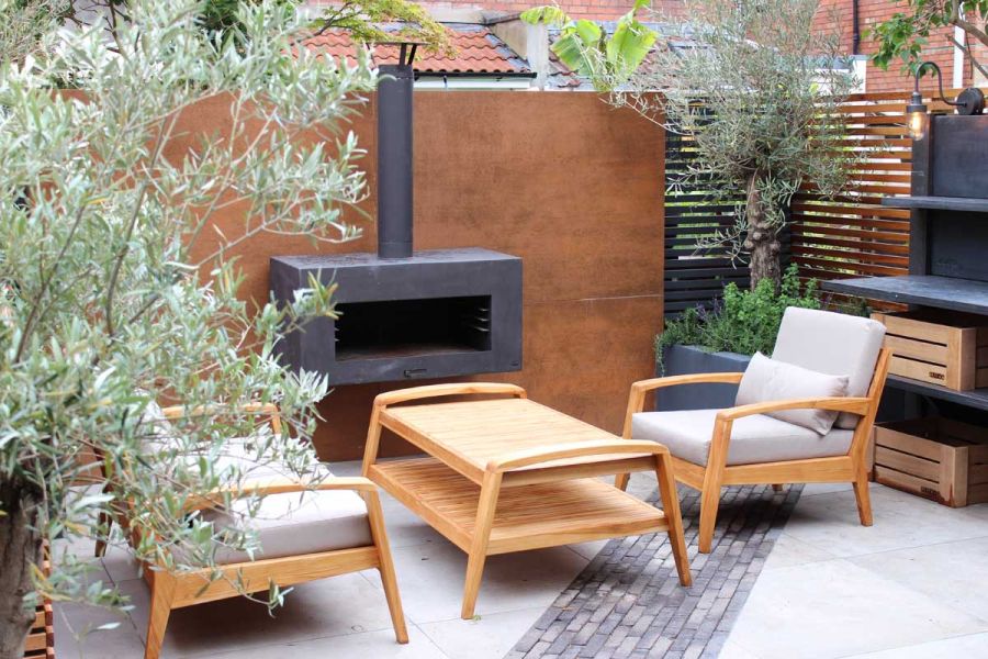 In garden corner, wooden lounge set next to black stove sit on paving bisected by diagonal strip of Silver Grey Multi clay pavers. 