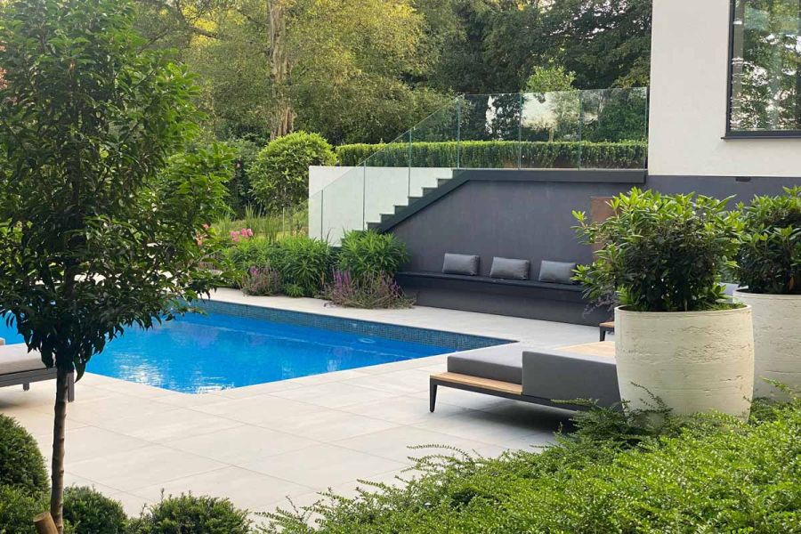 Large format Yard Porcelain around a swimming pool in modern garden, with green trees and shrubs. Chic poolside furniture in shot.