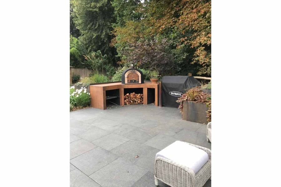 White-cushioned rattan footstool stands on patio of Black granite paving slabs, opposite bench with pizza over and log pile.