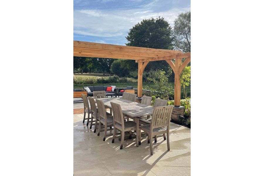 Ten seater wooden table and chairs sit under wooden pergola on a golden stone porcelain paving patio, lounge area at back.