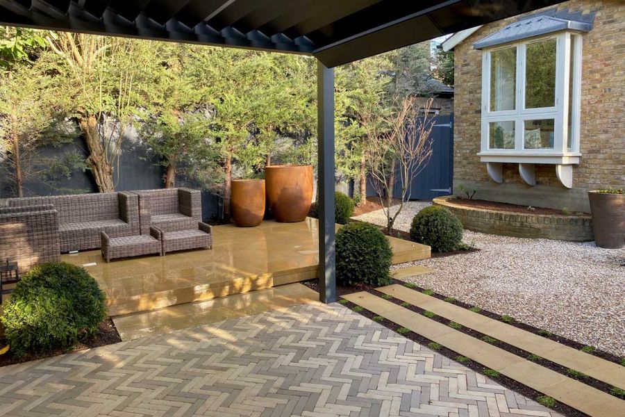 Pergola sits over Gromo Antica clay pavers at back of house, next to raised patio with furniture and pots. Design by Emma Griffin.