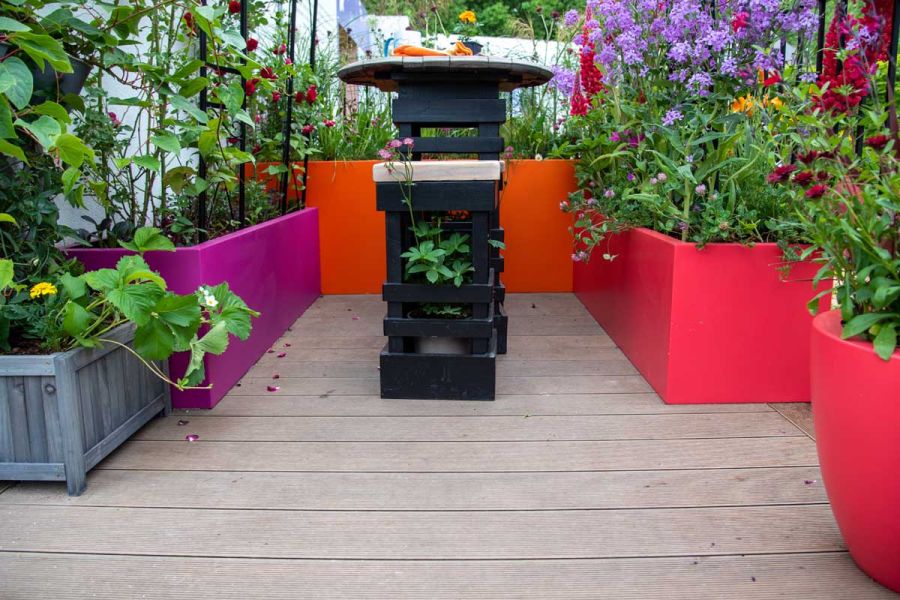 Black table and stool in reclaimed wood on Warm Teak Grooved DesignBoard composite decking, amid brightly coloured trough planters.