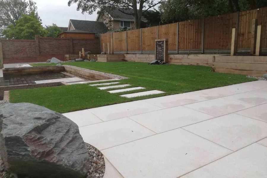 Large boulder sits by Florence White porcelain patio by Habitat Landscapes. Matching stepping stones cross lawn to sunken pond.