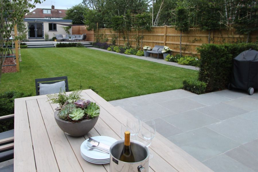 Large suburban garden with oblong lawn separating patio at back of house from Graphite Grey paved area with dining table.