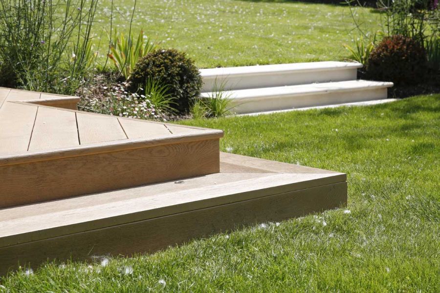 Sharp-angled corner of Golden Oak Millboard decking platform near 2 white steps, both set into lawn by Heartwood Garden Design.
