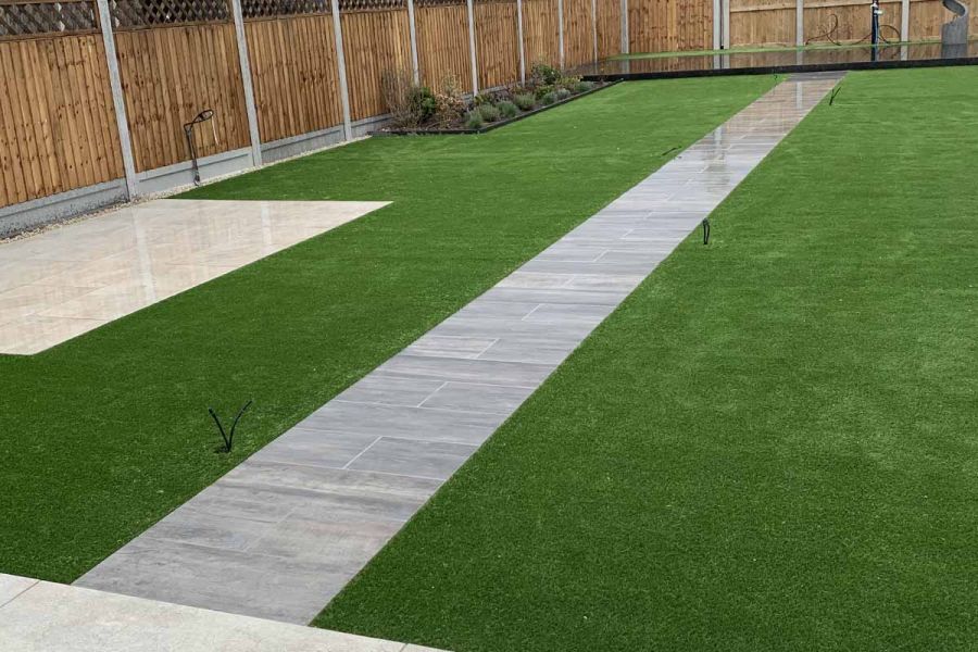 Thin strip of cinder porcelain paving leads up a wide lawn to a decking area and water feature with another small light patio on the right hand side.