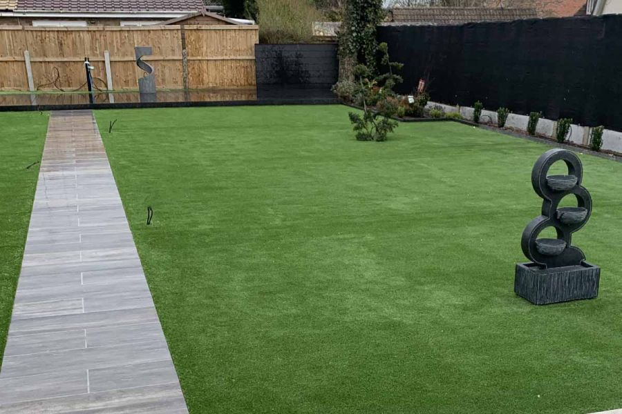 Thin strip of cinder porcelain paving leads up a wide lawn to a decking area and water feature. Plant bed alongside fence.