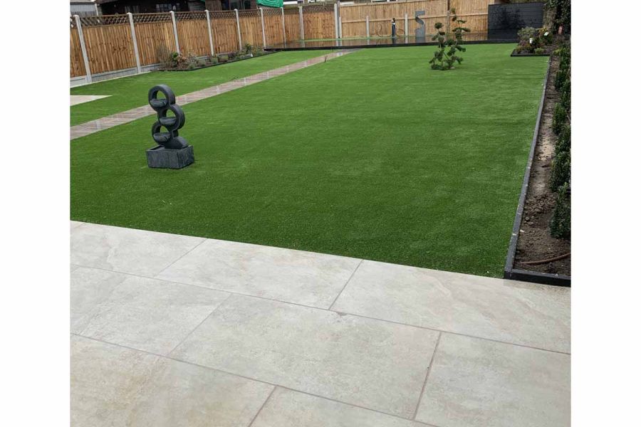 Low shot of Silver Grey Porcelain Paving in large garden with grass and flowerbeds with high fences surrounding.