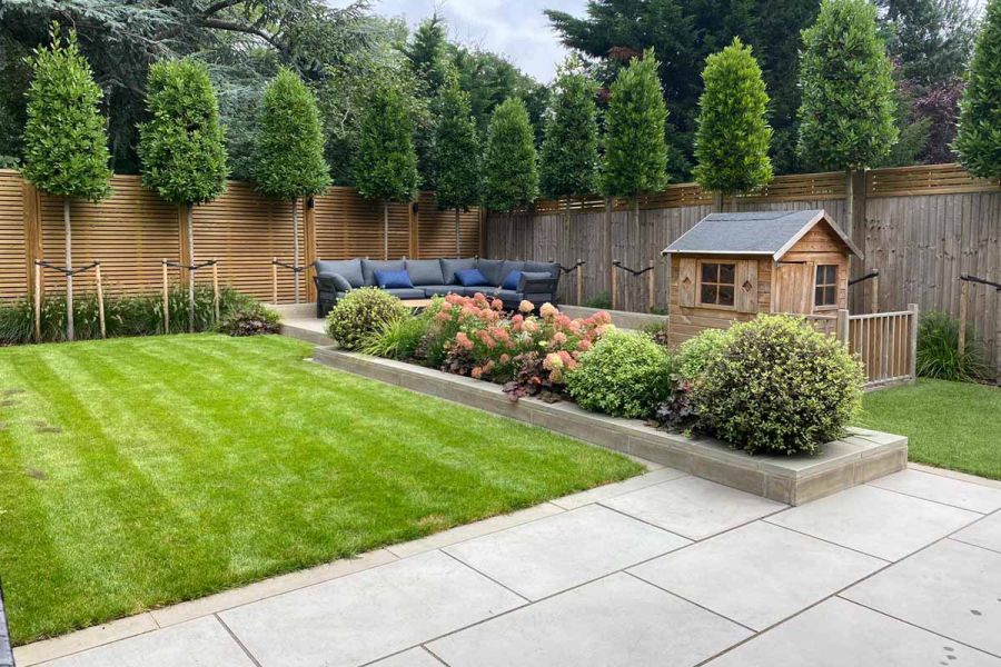 Fenced garden with lawn, corner sofa and Wendy house has shrub-filled raised bed topped with Heath Smooth Sandstone wall coping.