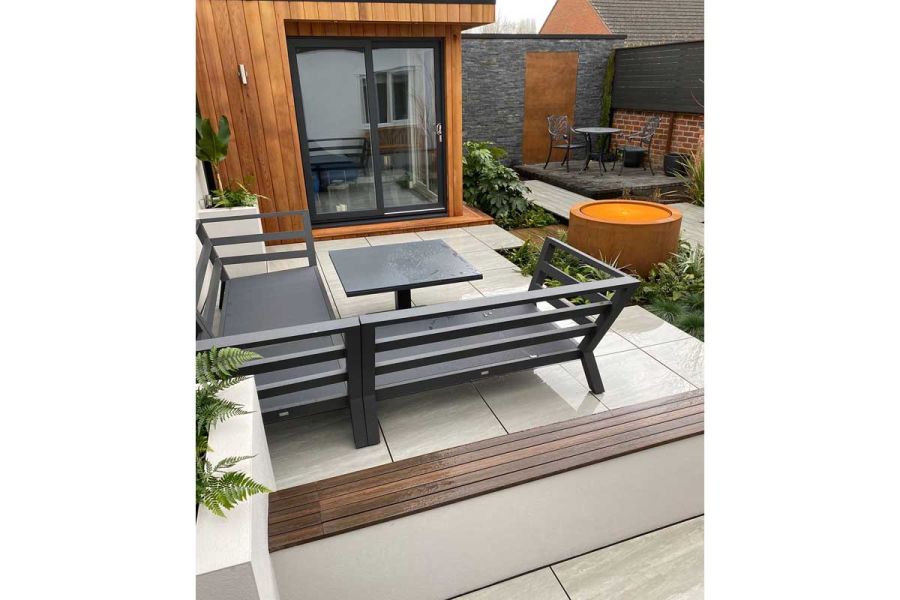 Metal sofa and table sit on porcelain paving on patio dividing garden room from low wall topped with Chestnut composite battens.