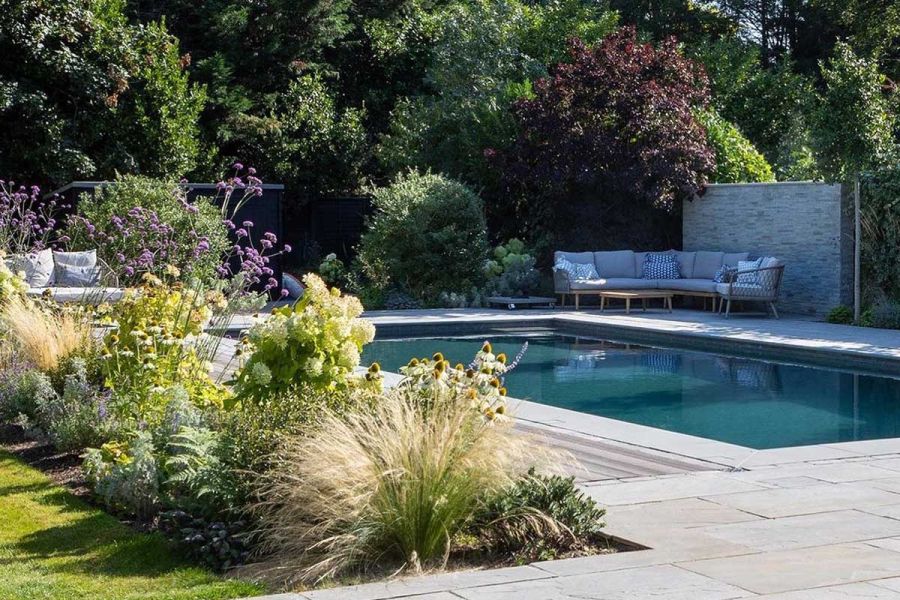 Oblong swimming pool with surround of decking and Kandla Grey Indian sandstone paving in corner of garden with mature trees behind.