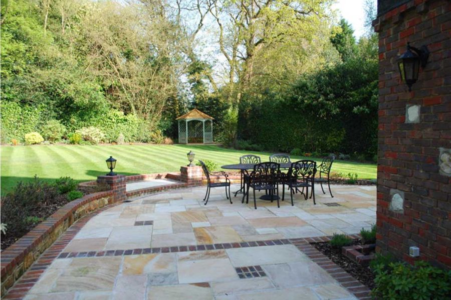 Large Mint Indian Sandstone patio wraps around corner of house. Paver-edged step up to lawn with summer house and mature trees.