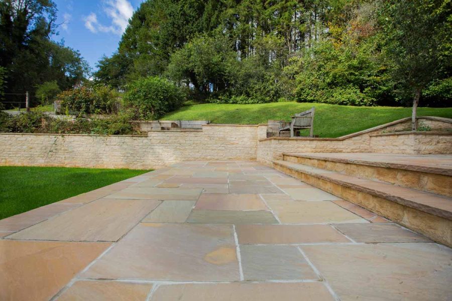 Wide steps separate 2 levels of Camel Dust Indian sandstone patio. Grass slope rises from behind retaining wall on far side.