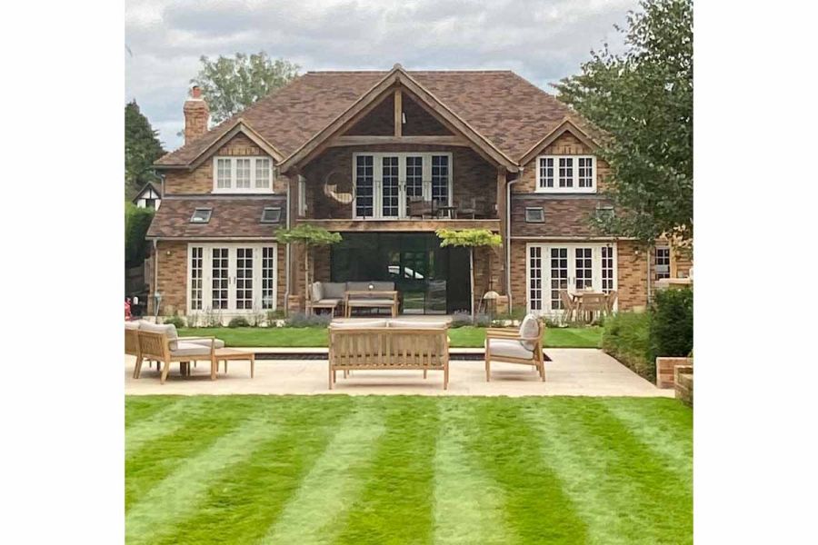 A picturesque view of large wooden house surrounded by lush lawns, with small patio paved in Florence Beige porcelain, adding warmth and elegance.