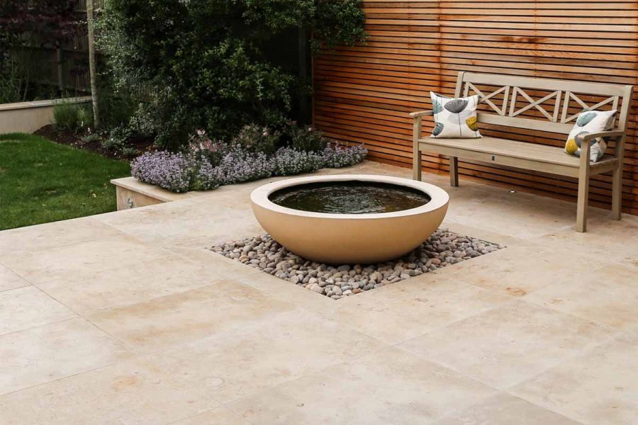 Shallow bowl-shaped pond sits on square of pebbles set into patio of Jura Beige smooth limestone paving next to wooden bench.