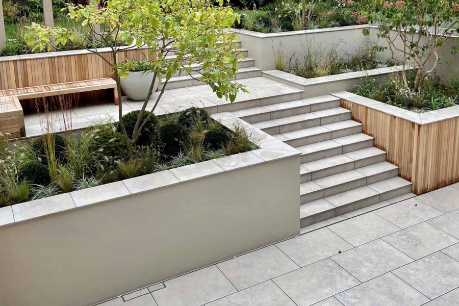 Jura grey porcelain garden steps rise from matching paving to terrace with wood benches, large white planter and rectangular bed.