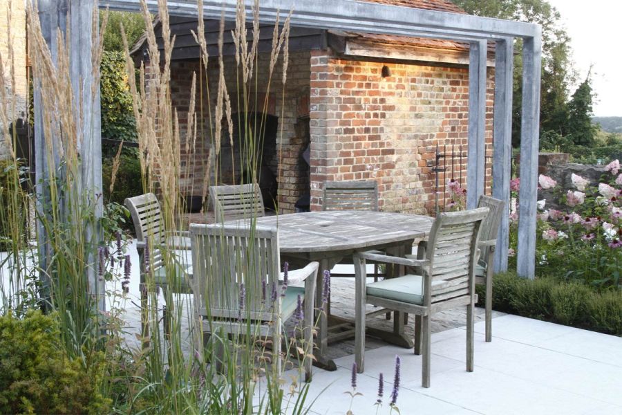 Wooden dining set on Jura grey limestone patio under metal arches. Brick shelter in background. Design by Greencube. Built by Land Design Partnership.
