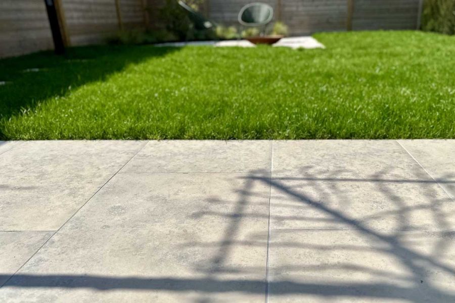 Shadows overlap a Close up view of Outdoor Tiles UK, the Jura Grey porcelain paving borders lush green grass.