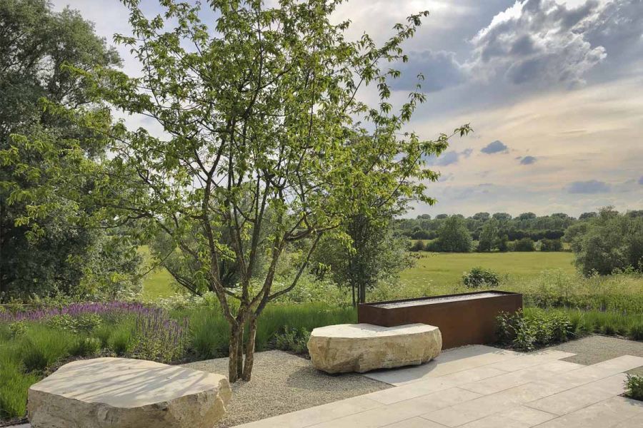 Jura Grey smooth limestone contemporary paving, with rusted water trough, stone boulders and prairie planting, overlooking fields.
