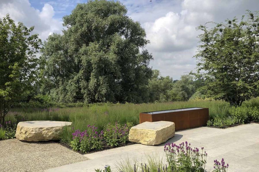 Jura Grey Smooth Limestone contemporary paving with gravel, water trough and flat boulders, edged by field with mature trees.