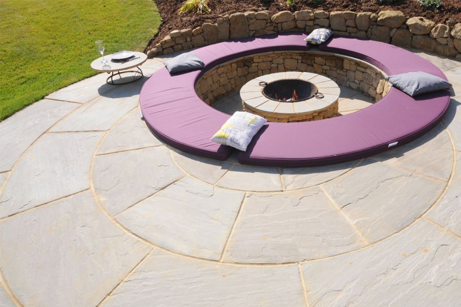 Bespoke natural paving circle in Kandla Grey sandstone offset from round sunken firepit with coping covered in purple fabric.