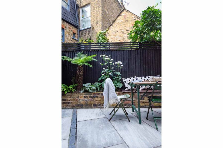 Metal chairs and table sit on Kandla Grey Indian sandstone paving in courtyard garden with brick raised bed lining tall fence.