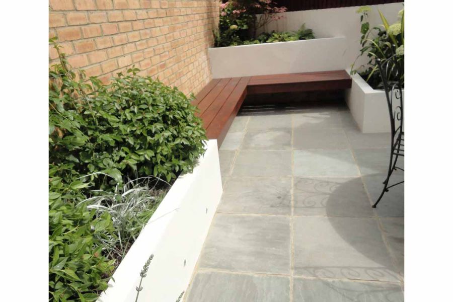 Corner of patio, next to tall brick wall, with white, raised beds and L-shaped inbuilt wood bench on Kandla Grey sandstone paving.