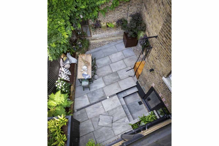 Aerial view of garden paved in Kandla Grey Indian Sandstone. Shrubs in raised beds and pots along edges. Steps down to back door.
