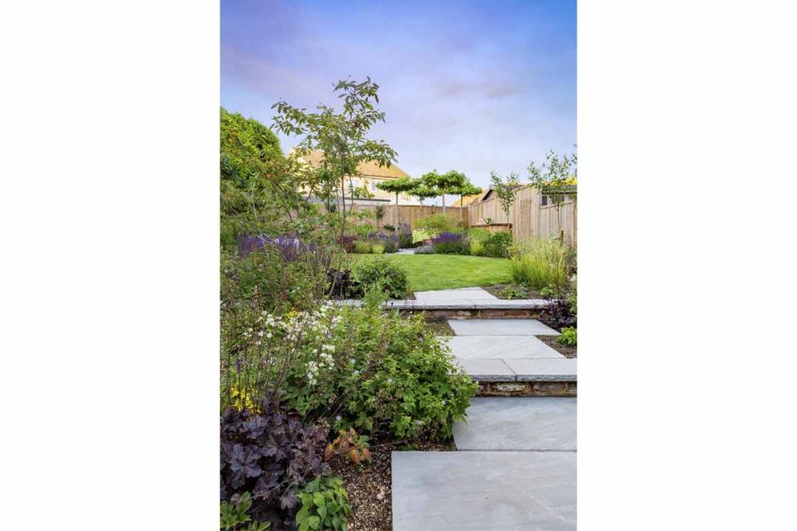 Staggered path of Kandla Grey Indian sandstone paving slabs leads up slope between flower beds to lawn, garden patio in far corner.