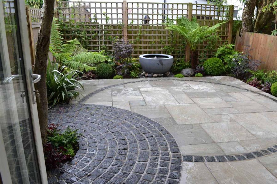 Small back garden with semi-circle of black granite setts cutting into Kandla Grey sandstone paving, edged with planted beds. 