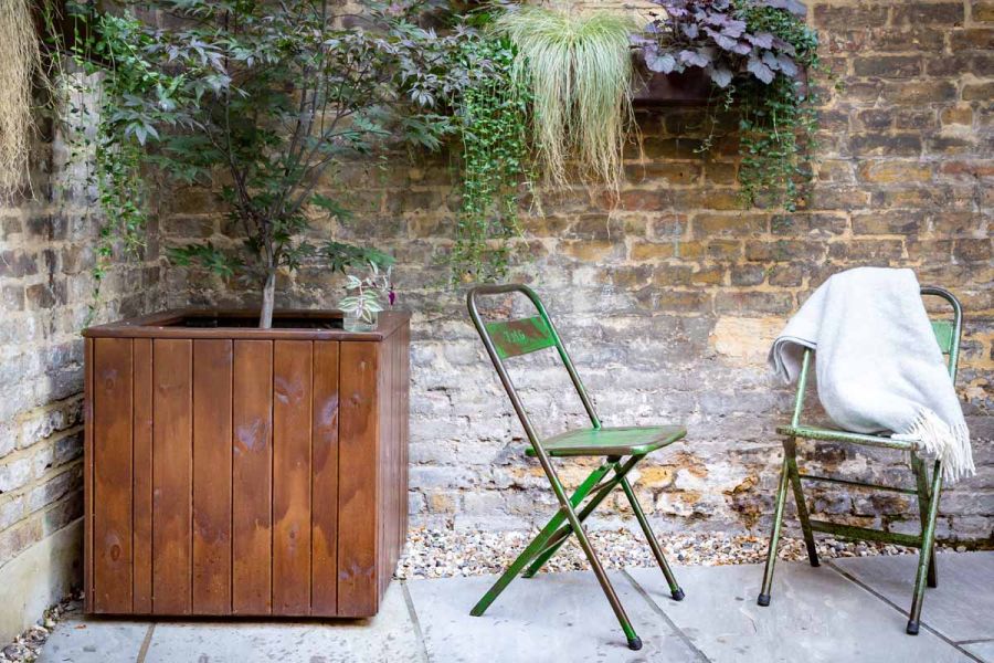 Distressed green folding metal chairs stand in in corner of Kandla Grey Indian sandstone garden patio next to large wooden planter.