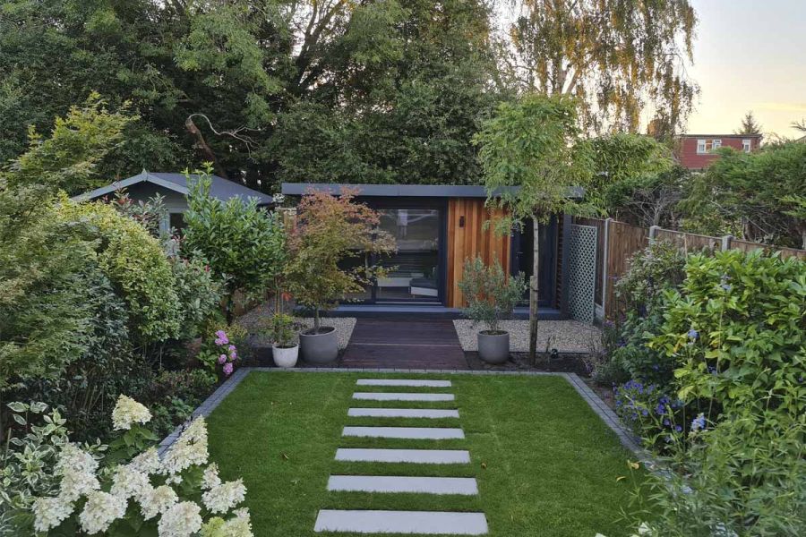 Spaced strips of Kandla Grey Porcelain paving lead across lawn to patio and summer house, past leafy borders and trees in pots.