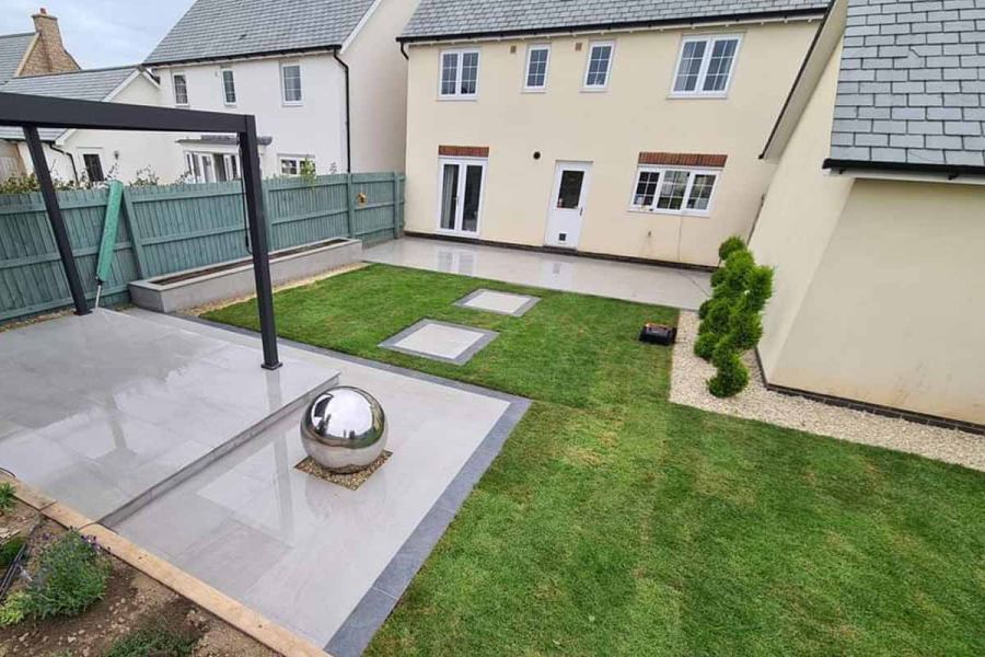 Silver ball sits on gravel square on 2-level patio of Kandla Grey porcelain, with dark plank edging in back garden with lawn.