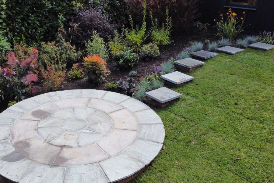 Next to long border, square stepping stones descend slope to Kandla Grey sandstone circle set which overlaps lawn and flower bed.