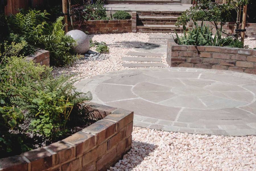 Kandla Grey sandstone paving circle set into gravel between curve-edged raised beds, with slab path leading to steps in background.