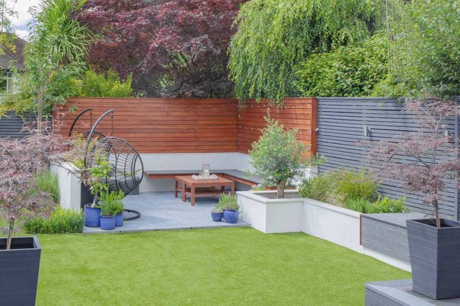 Kirkby Porcelain-paved corner enclosed by wooden cladding and wall. Planted blue pots next to raised beds and artificial lawn.