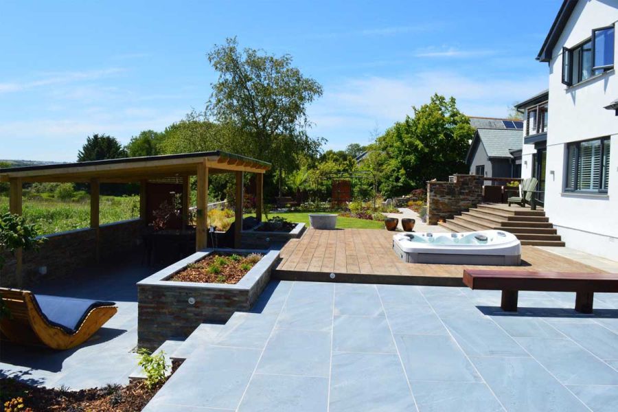 Large patio half paved in Kirkby Porcelain, half decked, with hot tub and steps down to large rocker chair and roofed pergola.