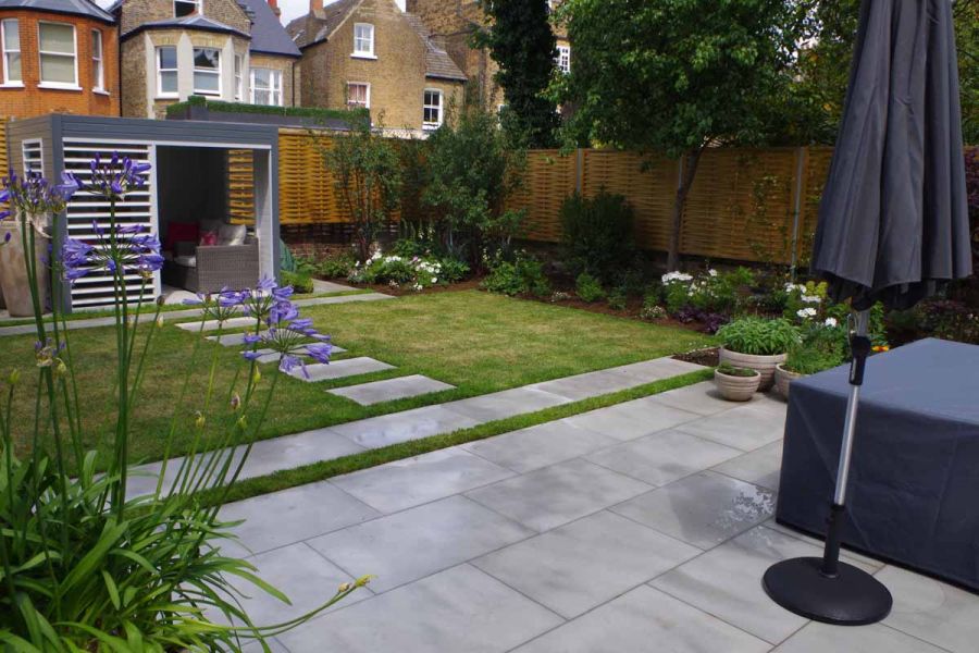 Contemporary Grey sawn sandstone stepping stones cross grass from house to matching paved area with freestanding parasol.